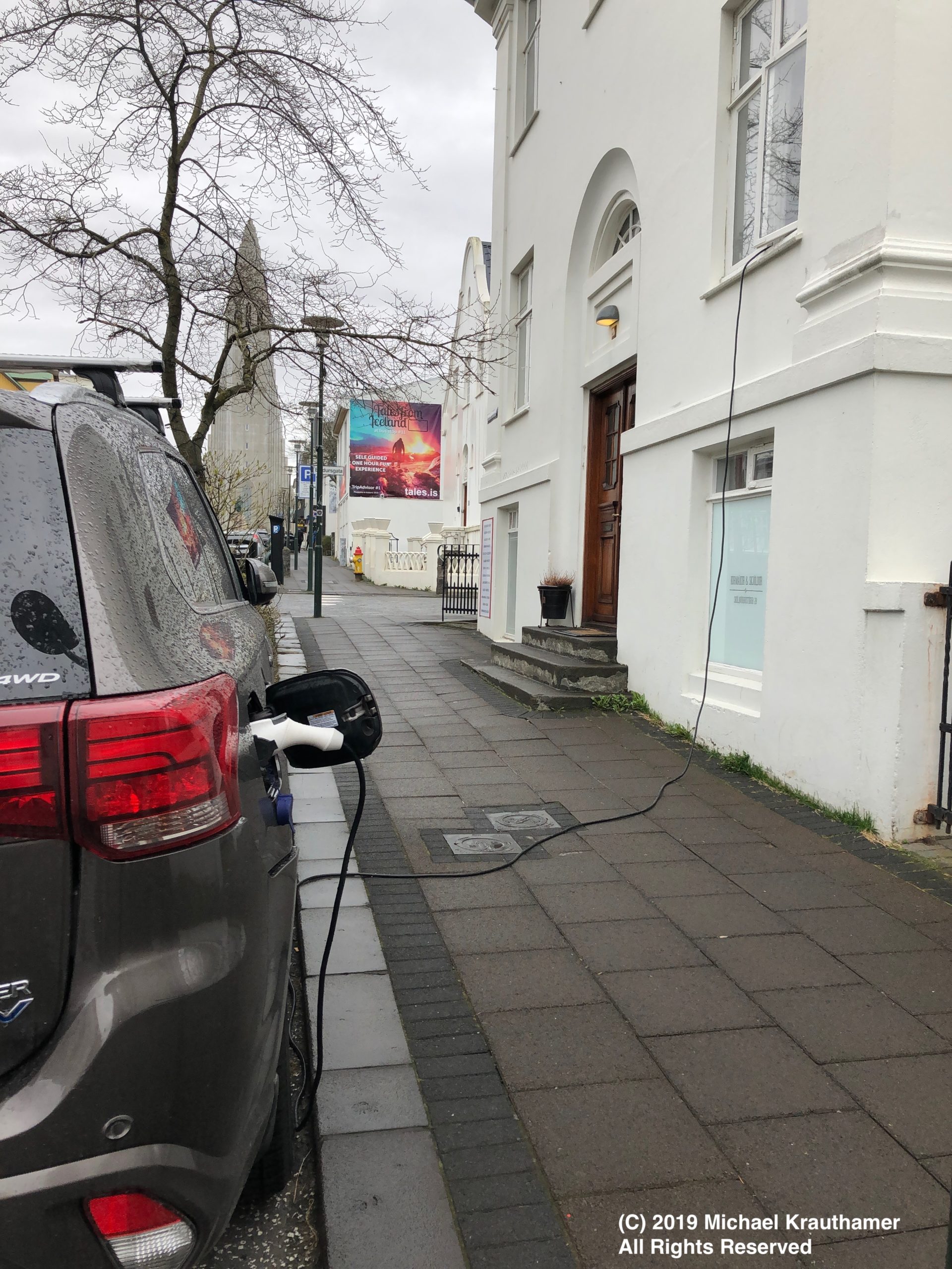 Photo of electric vehicle charging curbside over sidewalk and through window.